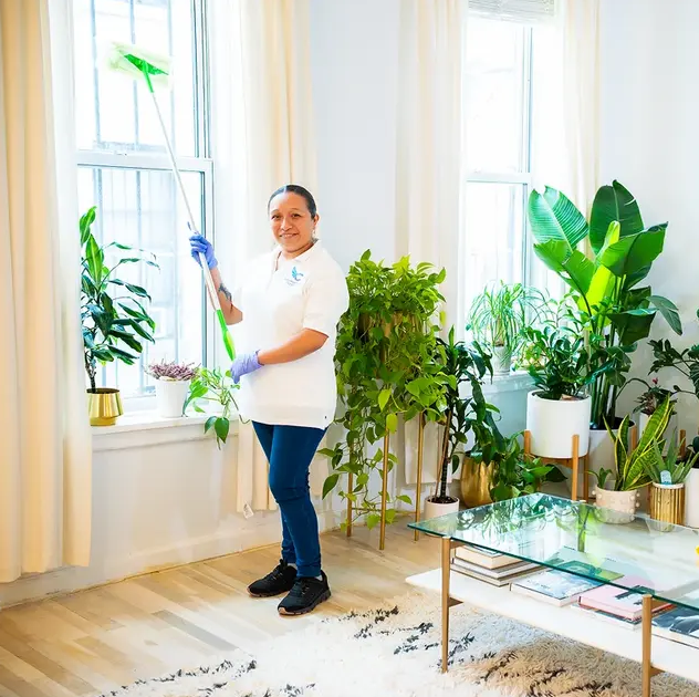 Smiling woman cleaning a window a brightly lit room filled with potted plants.